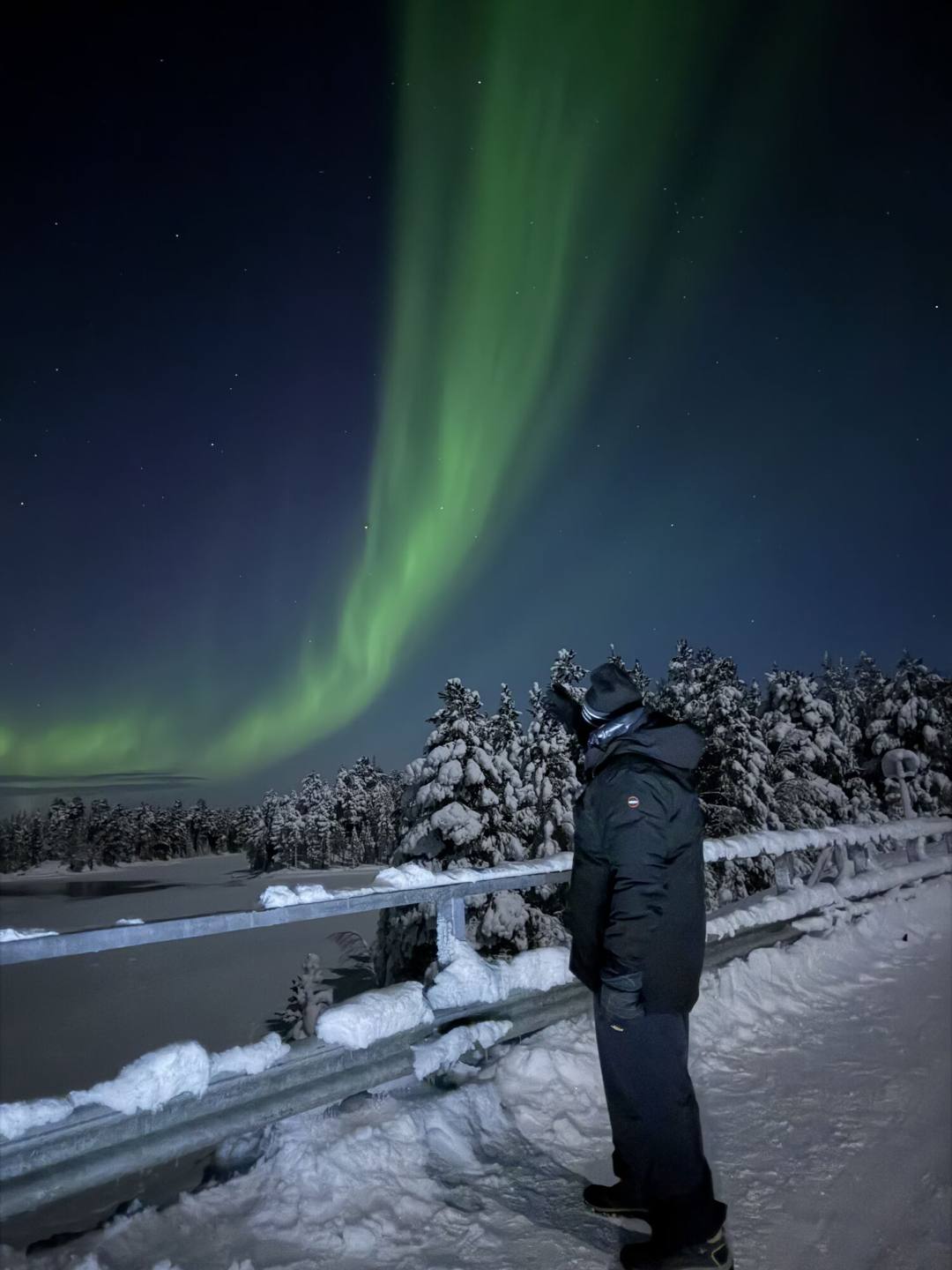 Guest at a snowy bridge railing looking up at a green aurora