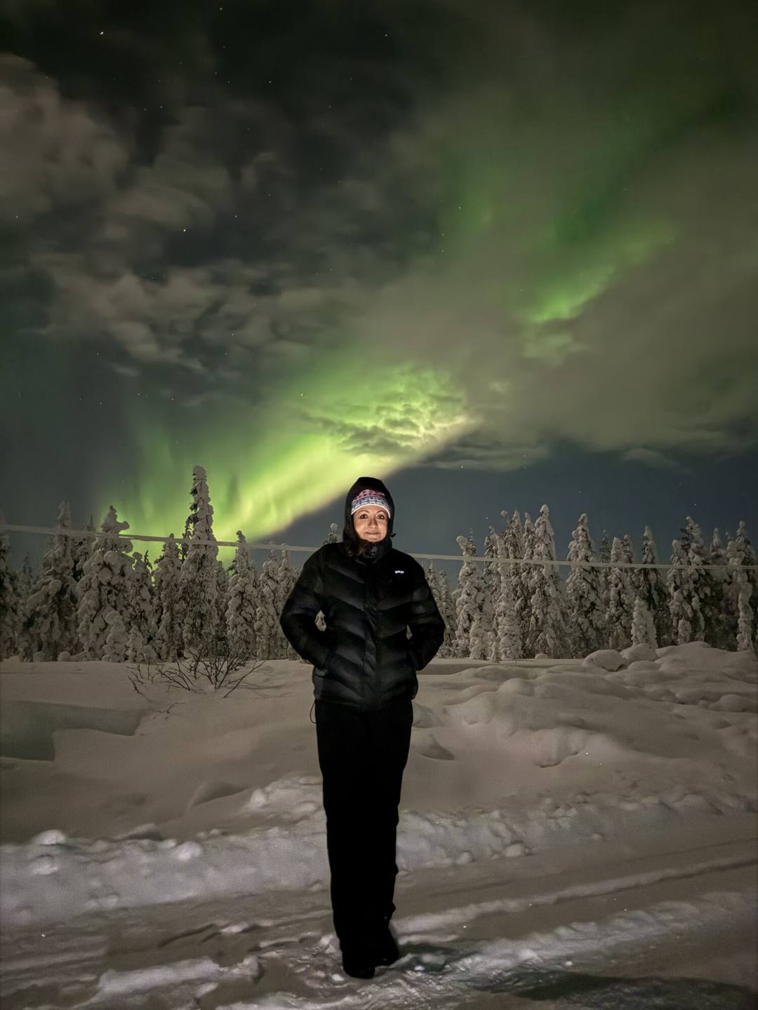 Guest in a black jacket standing in snow under a green aurora