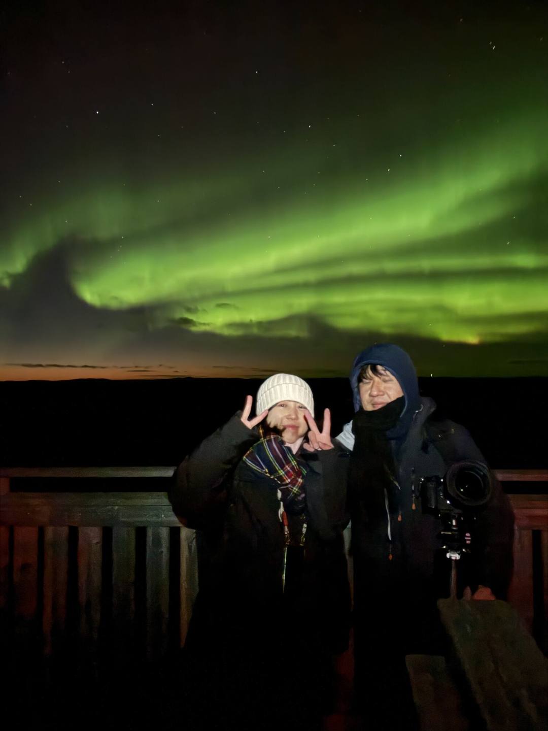 Two guests on a wooden deck under green Northern Lights
