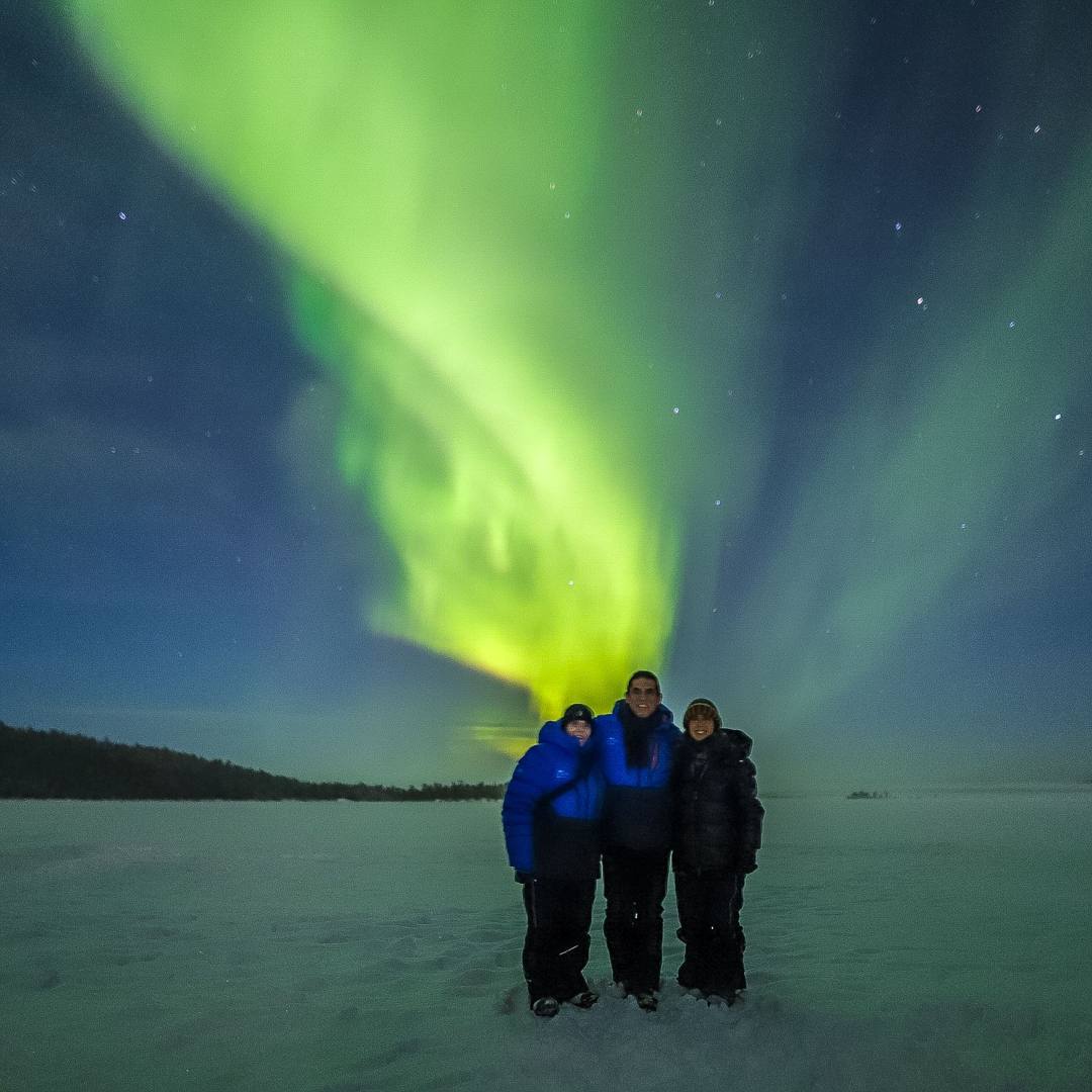 Three guests standing together on a snowy field under a bright Northern Lights corona