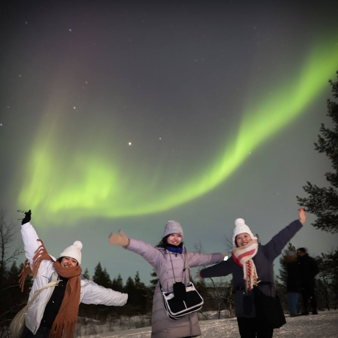 Three guests in winter clothing raising their arms under green Northern Lights