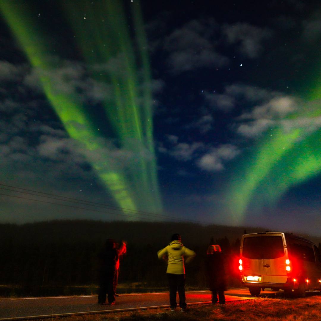Guests standing by a van on a roadside watching bright green Northern Lights