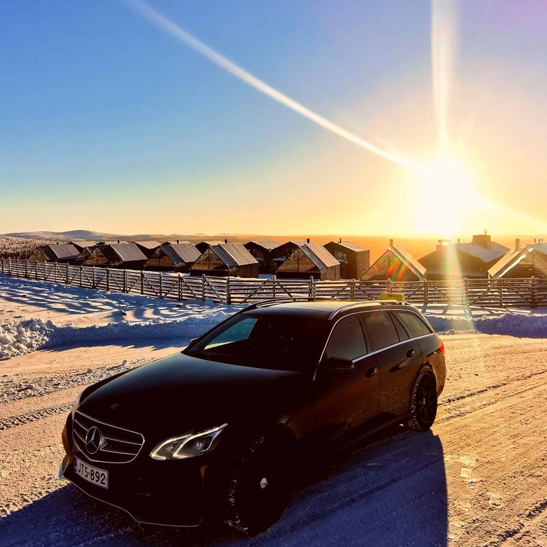 Black Mercedes-Benz estate car on a snowy road at sunset, with cabins in the background